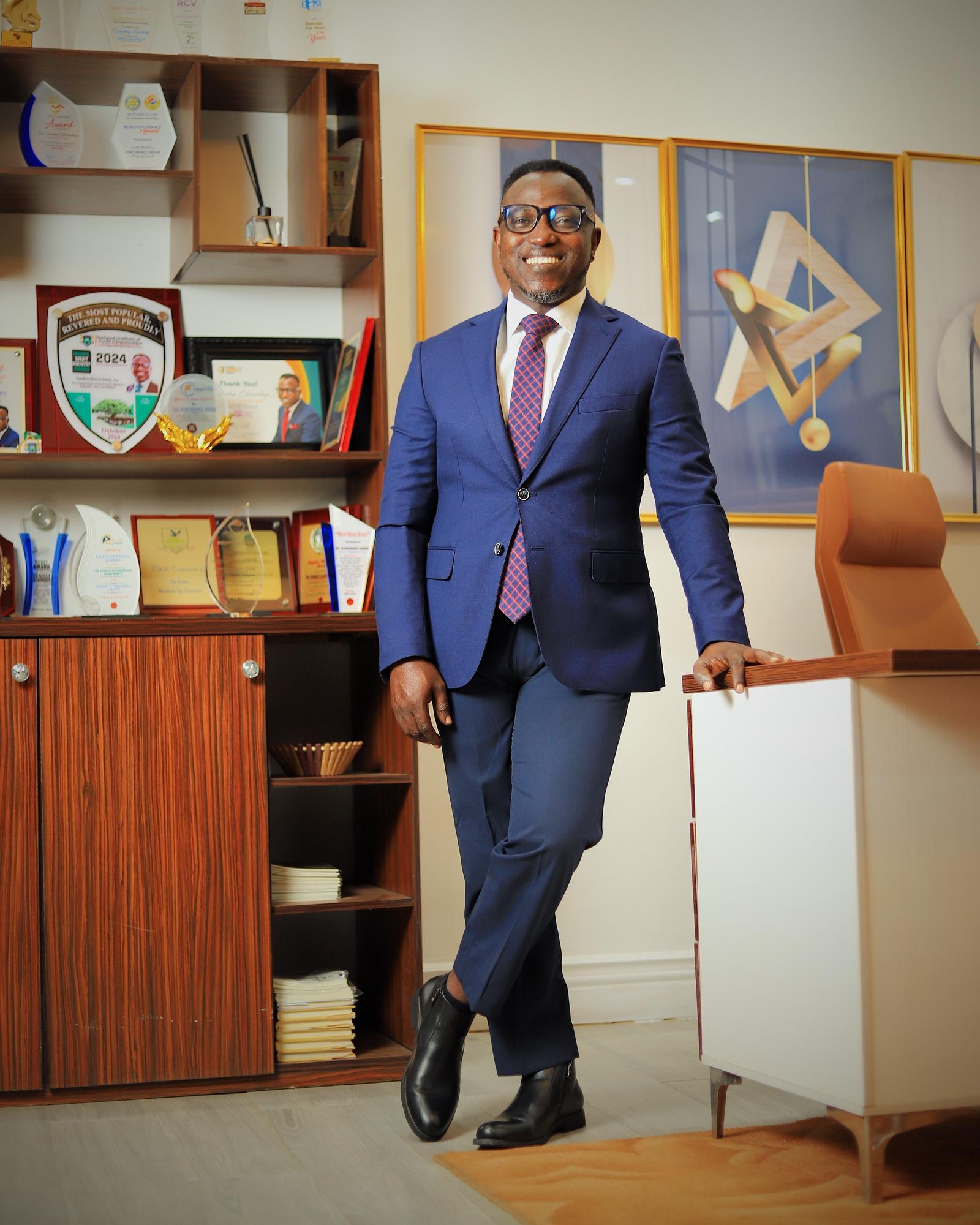 Dr. Sunday Olorunsheyi in a blue suit, leaning on a desk in an office lined with awards
